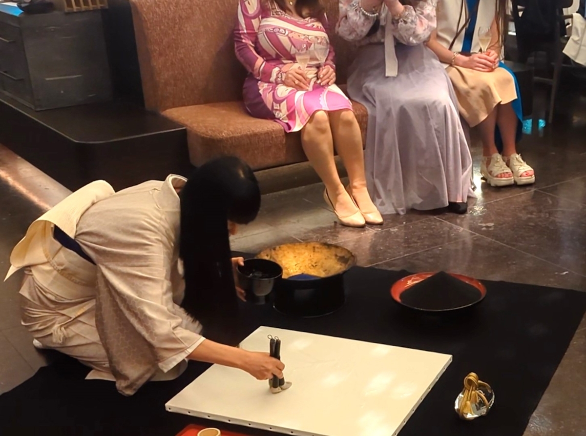 着物姿の松井ユカ氏が白いパネルに透明な糊で虎の一文字を書く