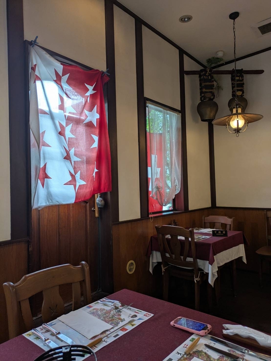 ル・シャレーの店内。壁に赤地に白い星のヴァレー州旗、奥の梁にカウベルが下がり、ワインレッドのクロスのテーブルが並ぶ