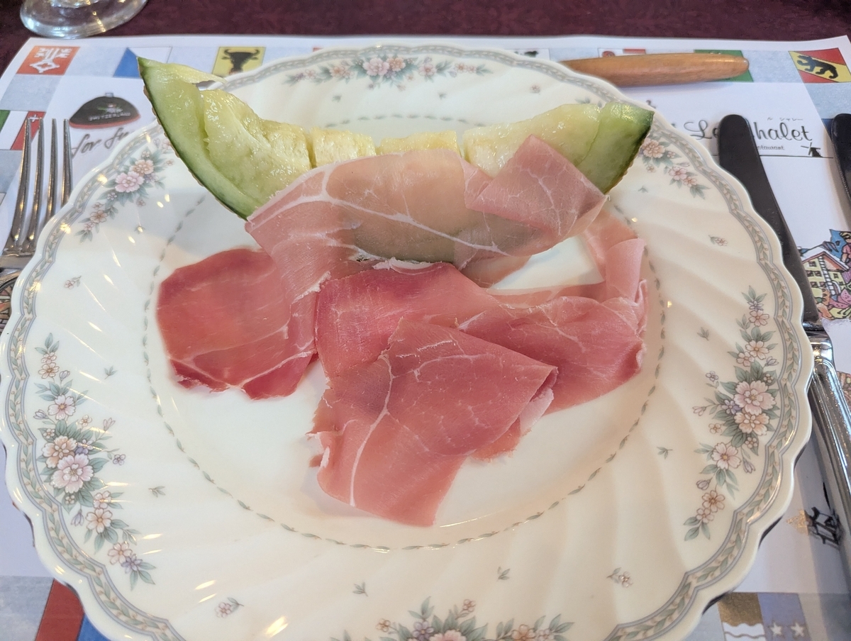 ル・シャレーのAコース前菜、生ハムとメロン。くし切りの緑肉メロンに薄切りの生ハムが掛かった花柄の皿
