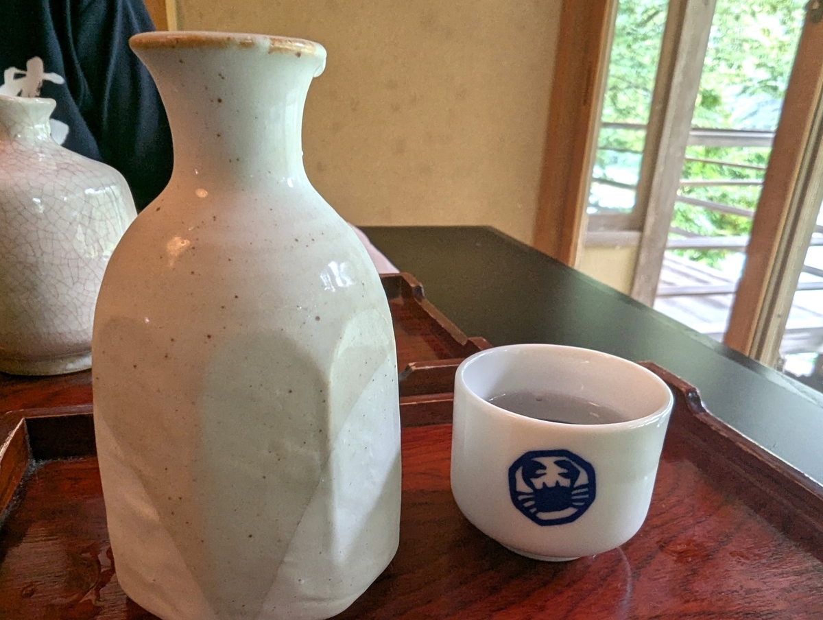 澤乃井の蟹の紋が入った猪口と白い徳利。窓の外に緑が見える