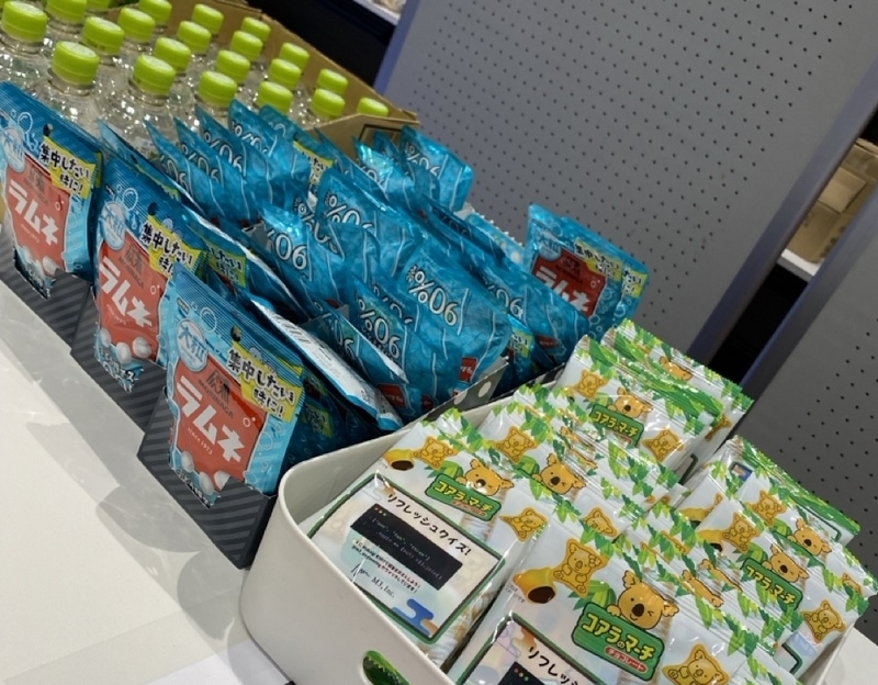 リフレッシュメントブースの机の上に、コアラのマーチ、ラムネ、水、お茶がたくさん並んでいる