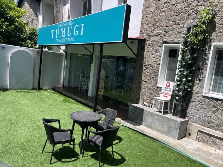 TUMUGIの店頭。水色の看板にJAPANESE STYLE TUMUGI CAFE &amp; SOFTCREAMの文字、人工芝のテラス席とラタン調のテーブル