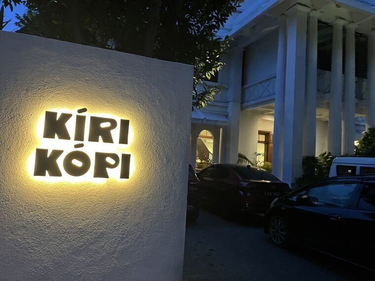 夜のKiri Kopiの看板。白い壁に立体文字のKIRI KOPIが電球色で照らされ、奥に白い列柱のコロニアル様式の建物