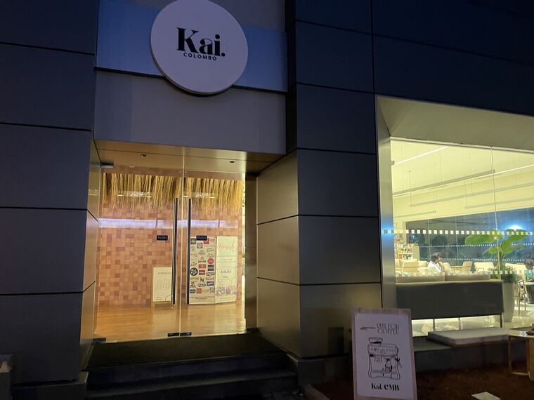 Kai. CMBの入口。丸い看板にKai. COLOMBO、ガラス扉の奥に藁を垂らした装飾とピンクのタイル壁