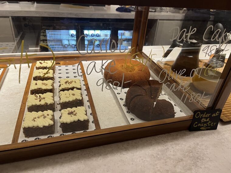 ケーキケースのガラスに手書きのラベル。Carrot Cake (GF) 980、Date Cake 980、Olive oil cake 1180の表記