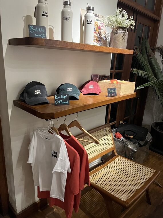 店内の物販棚。上段にKIRI KOPIロゴのボトル3本、下段に黒・青緑・ピンクのキャップとポストカードの木箱、下にロゴTシャツが吊られている