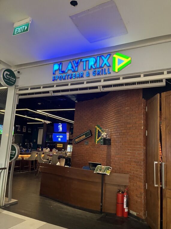 PLAYTRIX SPORTSBAR &amp; GRILLの青いネオン看板。入口の奥にレンガ壁とカウンター、スポーツ中継のTVが見える。左手前にIce-Cold Draughtの丸看板