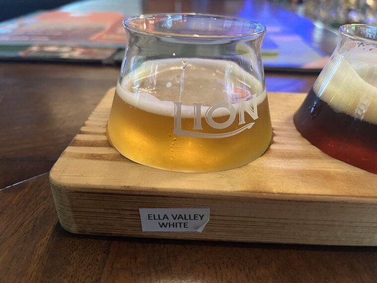 LIONロゴ入りグラスに淡い黄金色のビール。厚い白い泡。手前の木札にELLA VALLEY WHITE