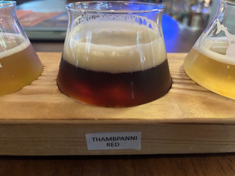 赤褐色から濃い琥珀色のビール。厚い白い泡。手前の木札にTHAMBPANNI RED