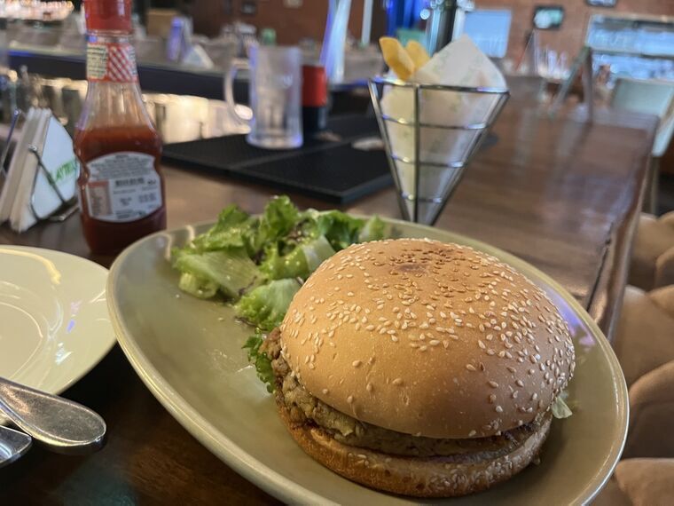 ゴマ付きバンズのハンバーガーと、オリーブオイルでマリネされたレタスのサラダが白い楕円皿に。奥に金属のカゴに入ったフライドポテトとケチャップ
