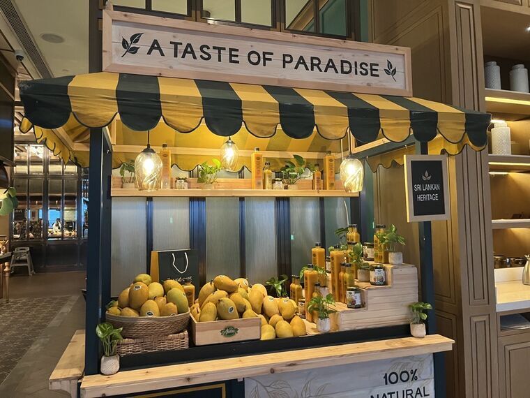 マンゴーのディスプレイ。A TASTE OF PARADISEの看板、SRI LANKAN HERITAGE、100% NATURALの文字。かごと木箱に生マンゴーが山盛り