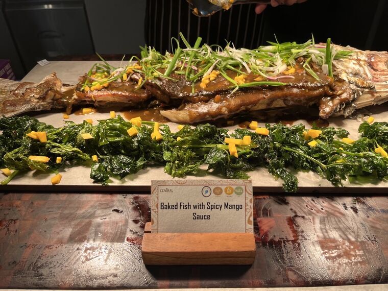 Baked Fish with Spicy Mango Sauceの名札と、丸ごと1尾の魚の姿焼き。白髪ねぎとマンゴーの角切りがトッピングされている
