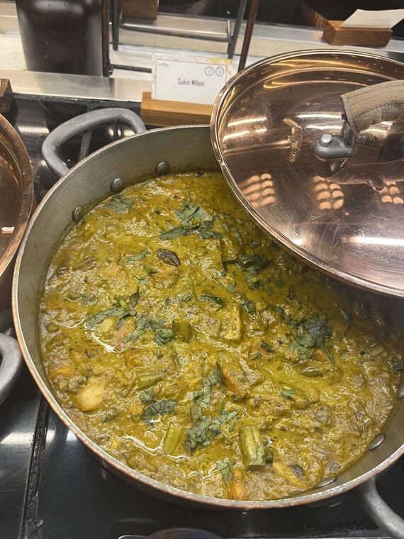 銅鍋の蓋を開けた緑色の野菜カレー。Subzi Miloniの名札が立っている
