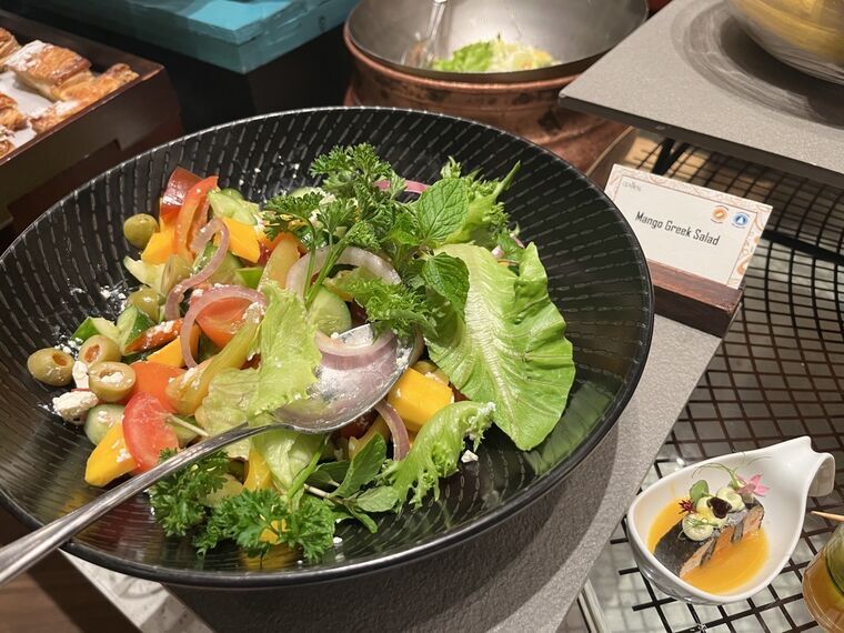 黒い器にレタス、トマト、玉ねぎ、マンゴーの角切り、オリーブ、フェタチーズ。Mango Greek Saladの名札