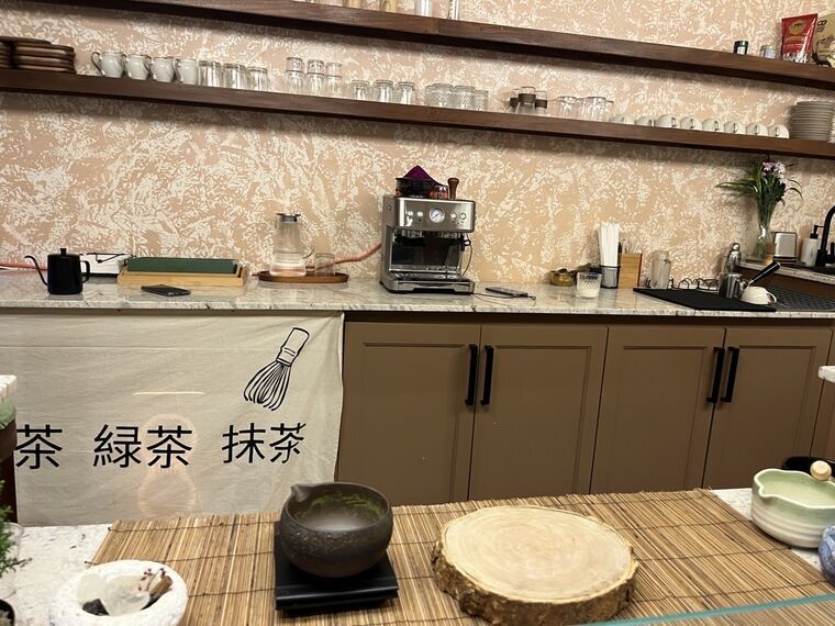 カウンター。背面棚にカップとグラスが並び、手前の布に日本語で茶・緑茶・抹茶の文字と茶筅のイラストが染め抜かれている