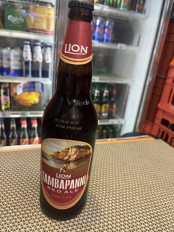 LION TAMBAPANNI RED ALEの瓶。赤いネックラベルにLIONロゴ、本体ラベルに崖と海のイラストとライオン、ABV 7.0%の表記。背景は酒売り場の冷蔵ケース