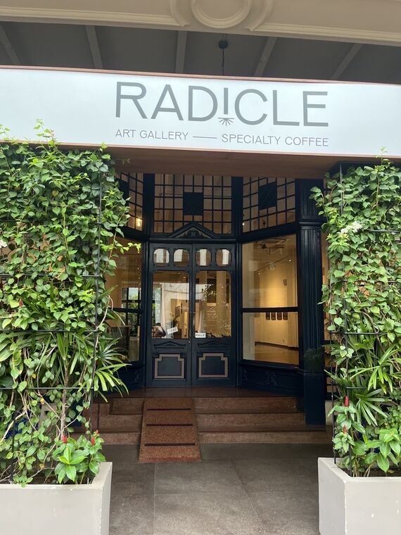 RADICLEの外観。白い看板にRADICLE、ART GALLERY &mdash; SPECIALTY COFFEEの文字。紺色の格子状の窓枠とガラス扉、両脇にツタが茂った鉢植え