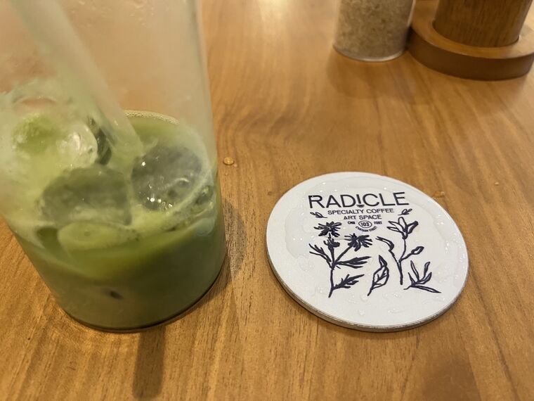 木製テーブルの上のRADICLEのコースター。RADICLE、SPECIALTY COFFEE ART SPACE、CMB 103 FORT CHATHAM STREETの文字と草花のイラスト。左に氷が溶けた抹茶ラテのグラス