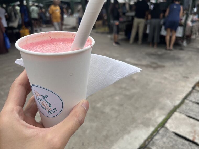 紙コップに入った鮮やかなピンク色のジュース。コップにZESTのロゴシール、紙ストローが刺さっている。背景はマーケットの通路