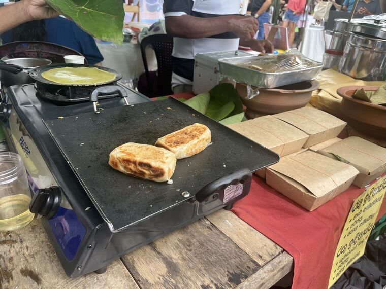 屋台の鉄板でマールロティ2つが焼かれている。奥のフライパンではホッパーの生地が焼かれ、右手に紙のテイクアウト容器が積まれている