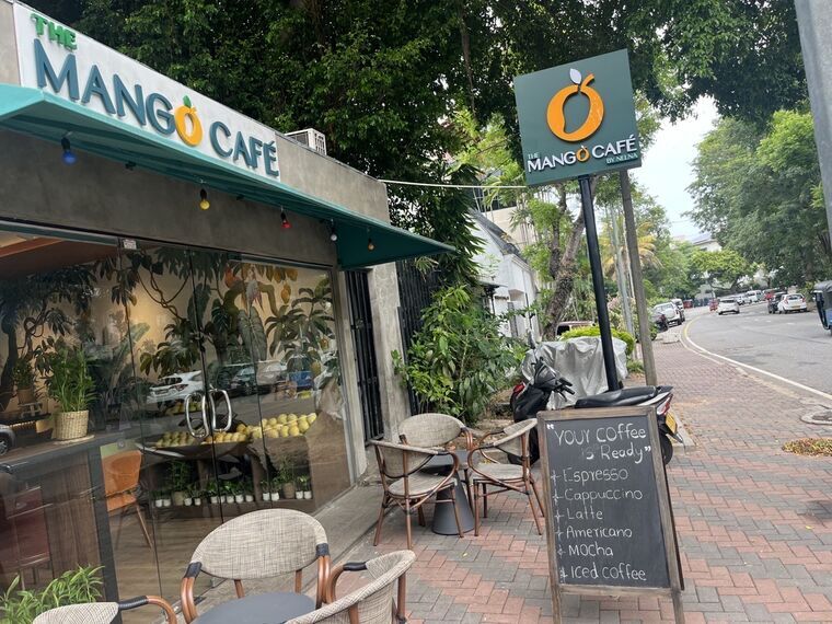 The Mango Cafe by Nelnaの外観。緑の庇にTHE MANGO CAFÉの立体文字とオレンジのマンゴーロゴ、道路脇に同じロゴの看板。テラス席と黒板メニューが並ぶ