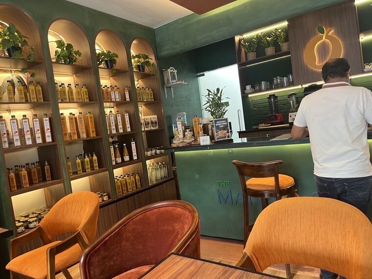 The Mango Cafe by Nelnaの店内。深緑の壁にアーチ型の棚が並び、オレンジや黄色の液体が入った瓶がびっしり陳列されている。オレンジとえんじの椅子、奥のカウンター上にマンゴーのネオンロゴ