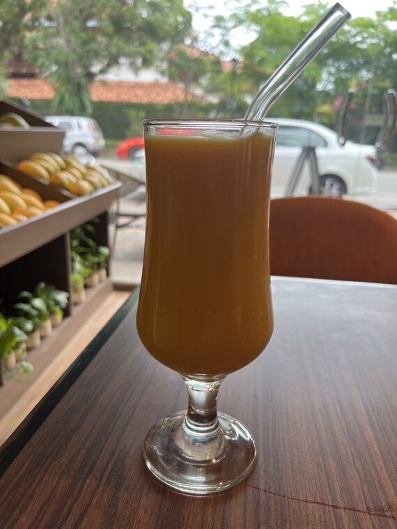 The Mango Cafe by Nelnaのマンゴージュース。脚付きのグラスにとろりとした濃いオレンジ色の液体が注がれ、ガラスストローが差してある。奥の窓際にマンゴーの陳列什器