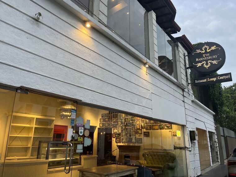 The Barnesburyの外観。夕暮れの白い壁の建物に、黒地に金文字の丸い看板でTHE BARNESBURY、その下にRestaurant Lounge Courtyardの表示。ガラス越しに額装写真が並ぶ壁と緑のソファが見える