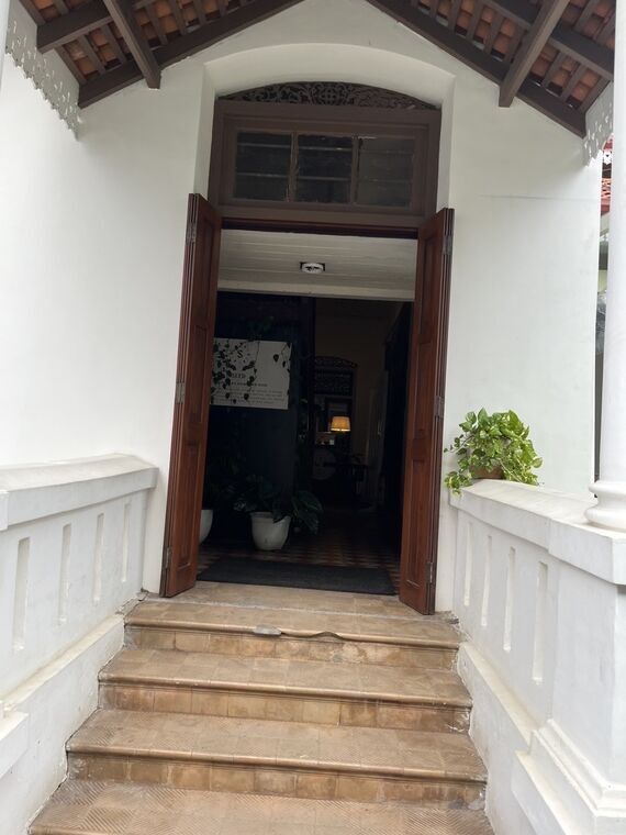 Seed Cafeの入口。瓦屋根の庇と白い手すりに挟まれたタイル張りの石段、その上に両開きの木製ドアが開いている。奥に市松模様の床と観葉植物、SEEDの案内板が見える