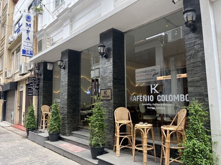 Kafenio Colomboの外観。黒い石張りの柱とガラス張りのファサード、KAFENIO COLOMBO、Speciality Coffee &amp; Matcha、Healthy Cuisinesの表示。ラタンのハイチェアとテラス席、鉢植えが並ぶ