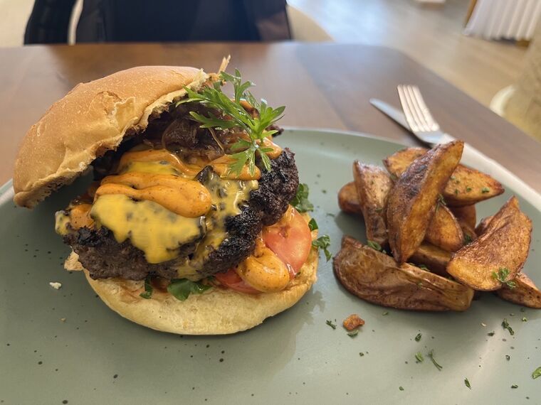 The Colombo Beef Burger。表面が黒く焦げた厚いビーフパティに溶けたチェダーチーズとオレンジ色のソース、キャラメリゼした玉ねぎ、トマト、葉物。上にパセリ。右に皮付きのポテトウェッジ