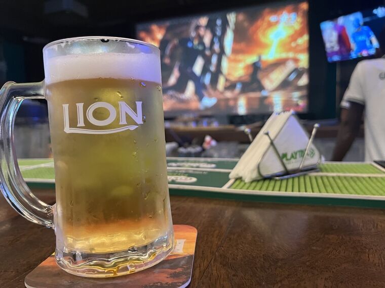 LIONロゴのジョッキに入った淡い黄色のビール。厚い白い泡。背景にクリケット中継の大型スクリーンとPLAYTRIXのナプキンスタンド