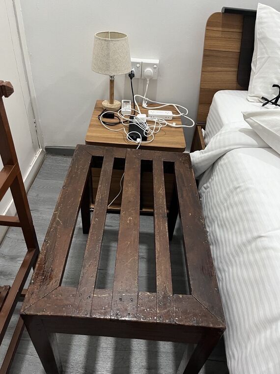 木製サイドテーブルに間接照明のランプ、エアコンのリモコン、モバイルバッテリー、電源タップと充電ケーブル。手前に木製の低いベンチ、右にベッド