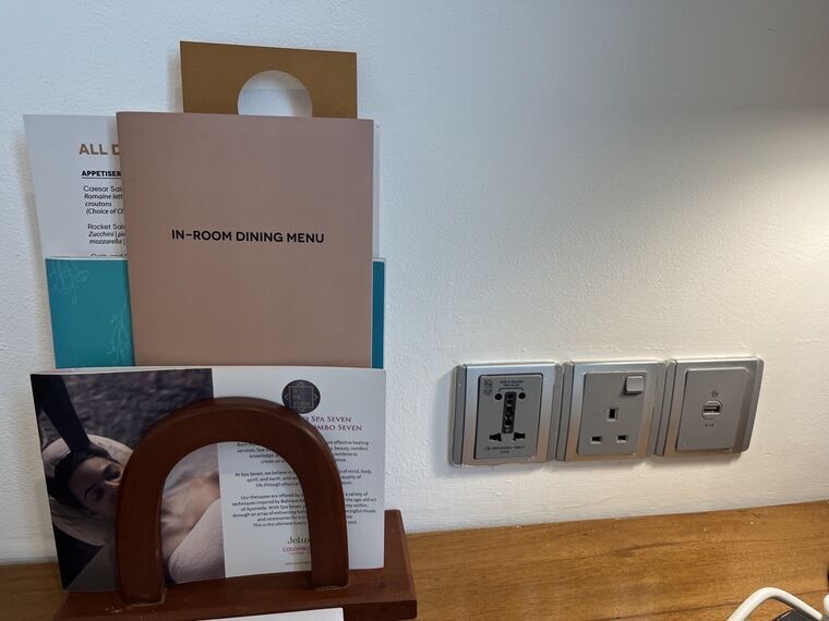 客室デスク横の壁にある3口のコンセント。丸ピン、BFタイプ、USBが1口ずつ。手前の木製ラックにIN-ROOM DINING MENUとスパの案内