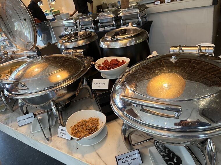 ビュッフェのスリランカ料理コーナー。銀色の保温容器が並び、STRING HOPPERS (v)、COCONUT SAMBOL (v)、LUNUMIRIS (v)、MILK RICE WITH GREEN GRAM (v)のネームプレートが立つ