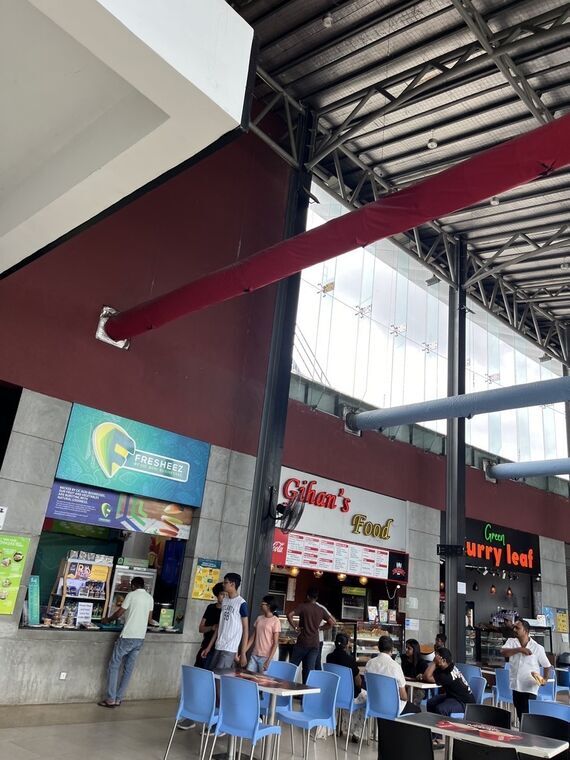フードコート。Gihan's Foodとgreen curry leafの看板、左にFRESHEEZのジューススタンド。水色の椅子とテーブルに客が座っている