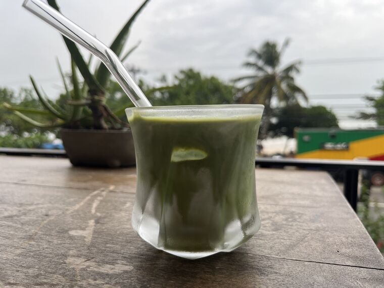 ガラスのタンブラーに入った緑色のアイス抹茶ラテ。ガラスのストローが刺さっている。木のテーブル、背景に鉢植えと道路、カラフルなトラック