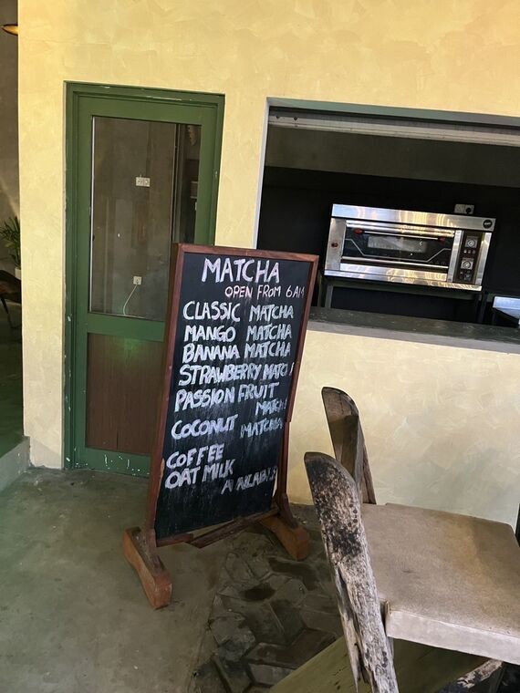 黒板にMATCHA、OPEN FROM 6AMと書かれ、CLASSIC MATCHA、MANGO MATCHA、BANANA MATCHA、STRAWBERRY MATCHA、PASSION FRUIT MATCHA、COCONUT MATCHA、COFFEE、OAT MILK AVAILABLEが並ぶ。背景に業務用オーブンと緑の扉