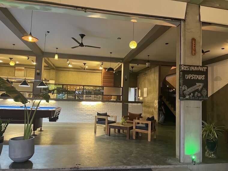 青いフェルトのビリヤード台とソファ、白いレンガのカウンター。右の柱にRESTAURANT UPSTAIRSと矢印の黒板。奥に階段