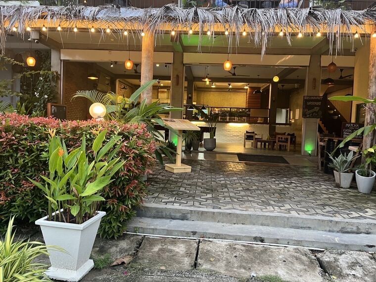 藁葺き屋根の軒下に電球のイルミネーション。奥に照明のついた店内、右手にMATCHAとRESTAURANT UPSTAIRSの黒板。手前に植栽と石畳