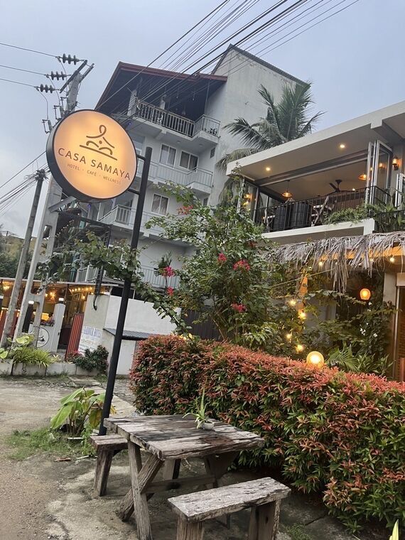 丸いオレンジ色の看板にCASA SAMAYA、HOTEL・CAFE・WELLNESSの文字。背後にバルコニー付きの白い宿泊棟、手前に木製のベンチテーブル