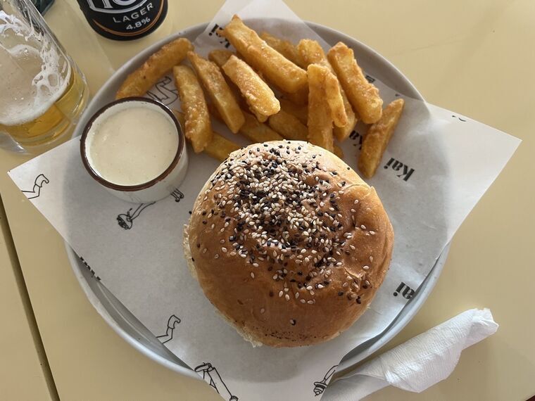 白い皿に胡麻のついたバンズのバーガー、太めのフライドポテト、白いディップの小鉢。手前にビールのグラス