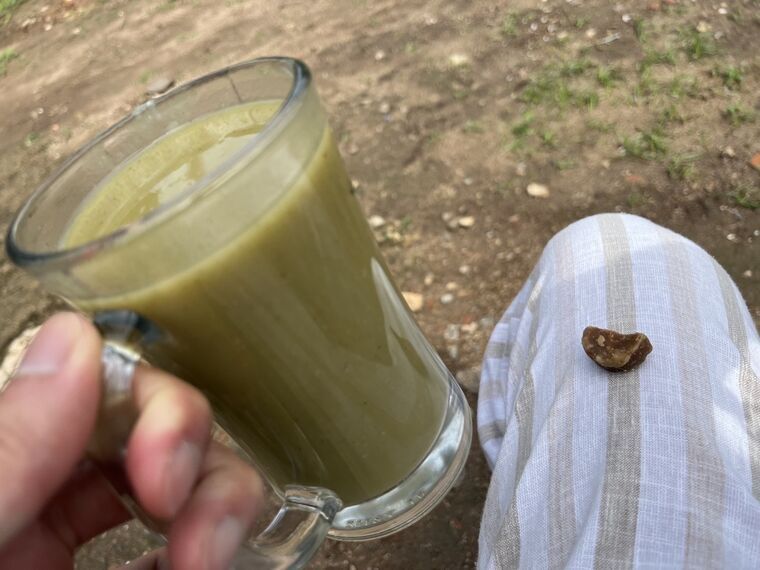 取っ手付きのグラスに入った緑がかった飲み物を手に持っている。膝の上の縞模様の布に黒糖のかけらがひとつ置かれている