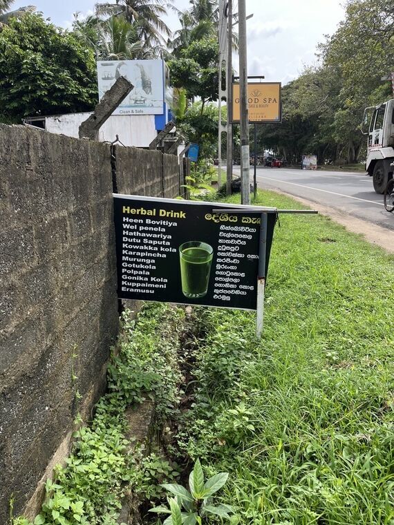 黒板にHerbal Drinkと書かれ、その下にHeen Bovitiya、Wel penela、Hathawariya、Karapincha、Murunga、Gotukola、Polpalaなどの薬草名がシンハラ文字と英字で並ぶ