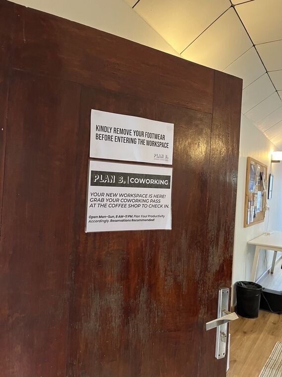 木製ドアに貼られた2枚の掲示。KINDLY REMOVE YOUR FOOTWEAR BEFORE ENTERING THE WORKSPACEと、PLAN B. COWORKINGの案内。Open Mon-Sun, 8 AM-11 PMの表記