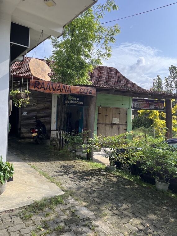 木彫りの「RAAVANA CAFE」看板が掛かる入口。赤瓦の屋根と緑色の木の扉、手前に石畳とバイク1台