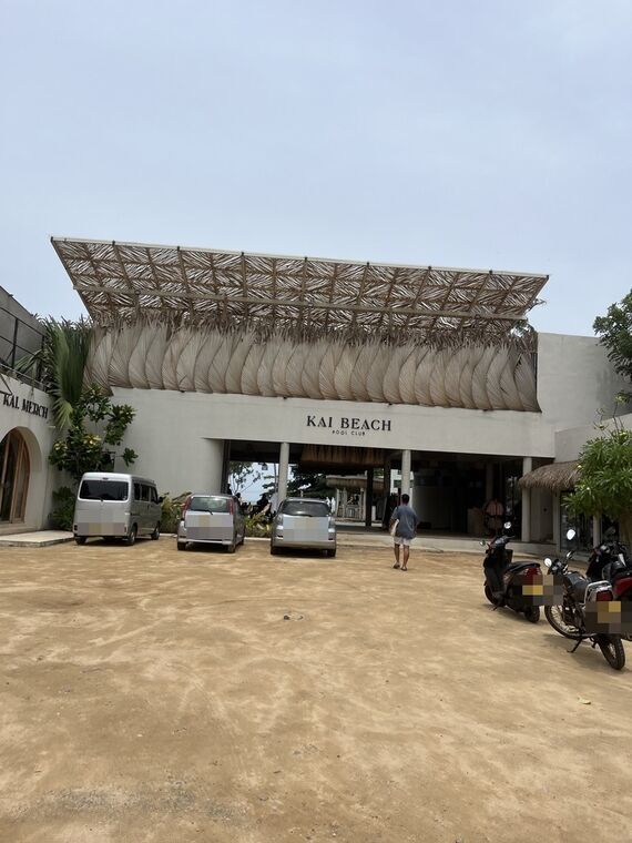 KAI BEACH POOL CLUBと書かれた建物の正面。椰子の葉を編んだ大きな庇、手前は砂地の駐車場に車とバイク。左にKAI MERCHの入口