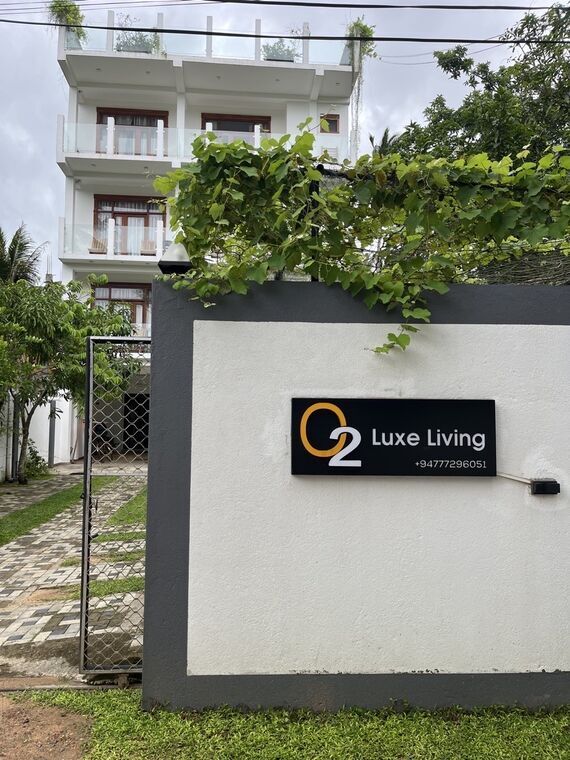 門の塀に設置された黒地の看板。O2 Luxe Livingの表記。背景に建物の外観と石畳のアプローチ