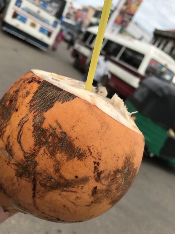黄色いストローが挿さったking coconutを手で持っている。背後にバスとトゥクトゥクが停まっている