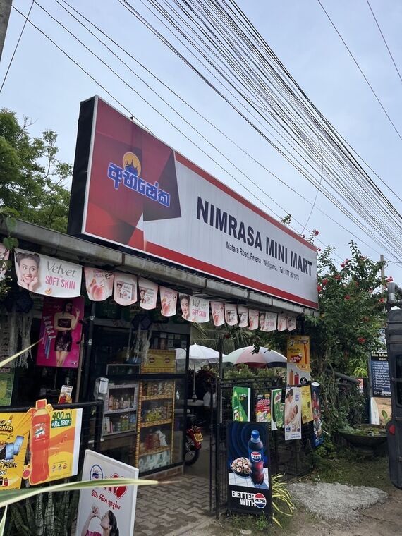 NIMRASA MINI MARTの看板。Matara Road, Pelena - Weligamaの住所表記、軒先の吊り下げ広告、店頭のPepsiの立て看板
